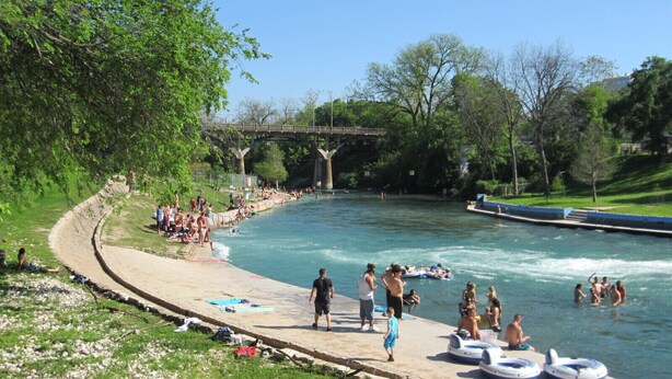 Zona para nadar a orillas del río, con un paseo marítimo de hormigón, balsas hinchables y gente disfrutando del agua.