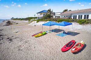 Ein Strand mit Kajaks und Sonnenschirmen, ein sandiger Uferstreifen und Gebäude im Hintergrund.