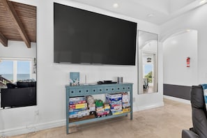Ein Wohnzimmer mit einem großen Flachbildfernseher an der Wand, einem hellblauen Konsolentisch mit Gesellschaftsspielen und Blick ins Freie durch ein Fenster.