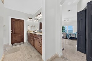 Un couloir avec une porte en bois, une salle de bain avec un meuble-lavabo et des miroirs, et un lit avec vue.