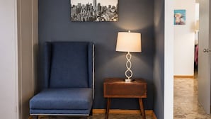 Um quarto com uma cadeira azul, uma lâmpada sobre uma mesinha de madeira e uma pintura com vista para a cidade na parede.