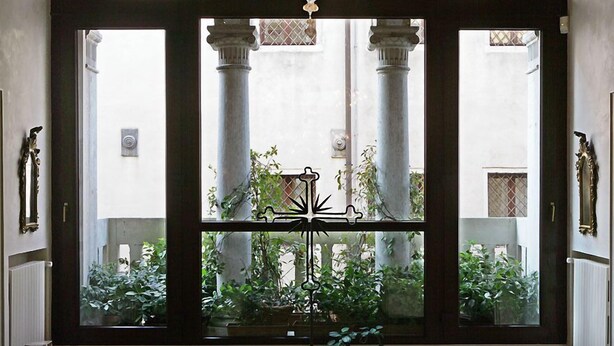 Blick durch ein Fenster mit schmiedeeisernem Gitter, Säulen und einem Kronleuchter.