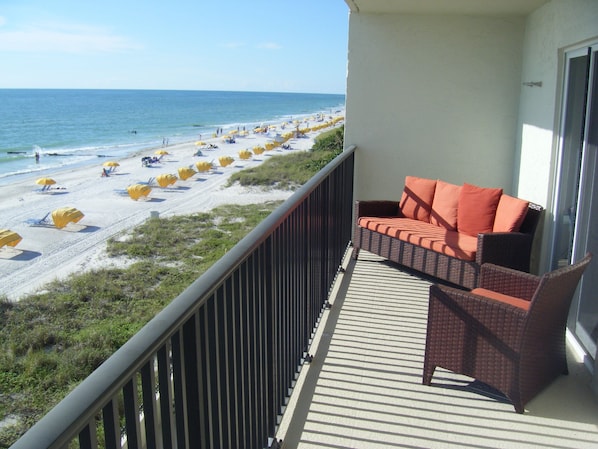 A balcony with a wicker sofa and orange cushions, overlooking a beach with yellow umbrellas and people. - Special Aug 29 to Sept 05, 2026 - Madeira Direct Beach Front Corner Condo (Madeira Beach)