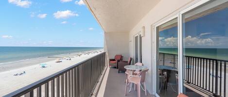 A balcony with a view of the beach, a table with chairs, and a wicker sofa.