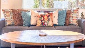 Un salón con un sofá decorado con cojines estampados, una mesa redonda y un ventanal con vistas a árboles.