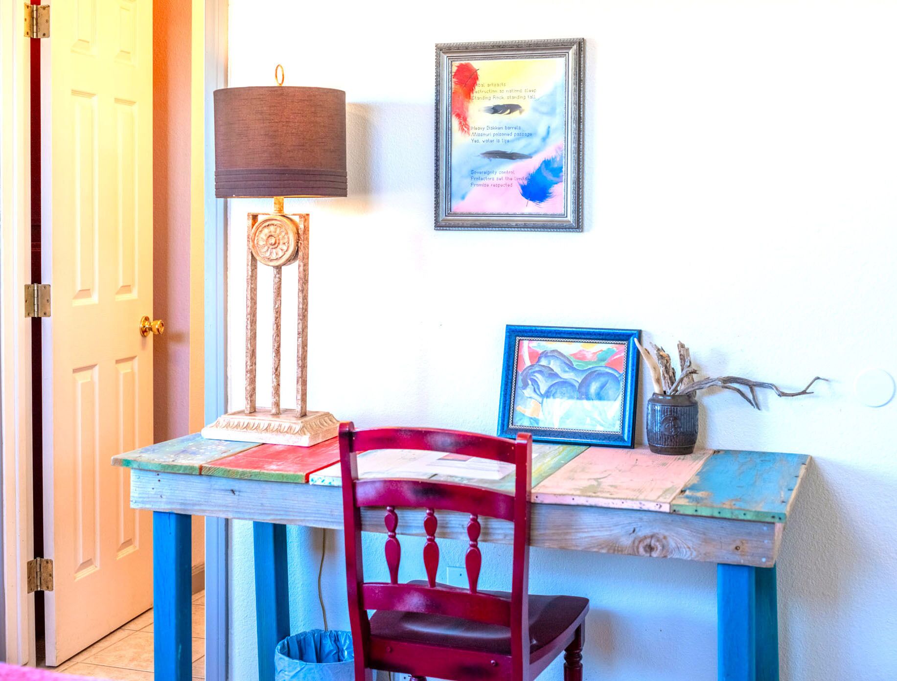 Una habitación con un escritorio de madera, una silla roja, una lámpara, un cuadro enmarcado y un florero con plantas secas.