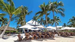 A beachside restaurant with outdoor seating, palm trees, and umbrellas.