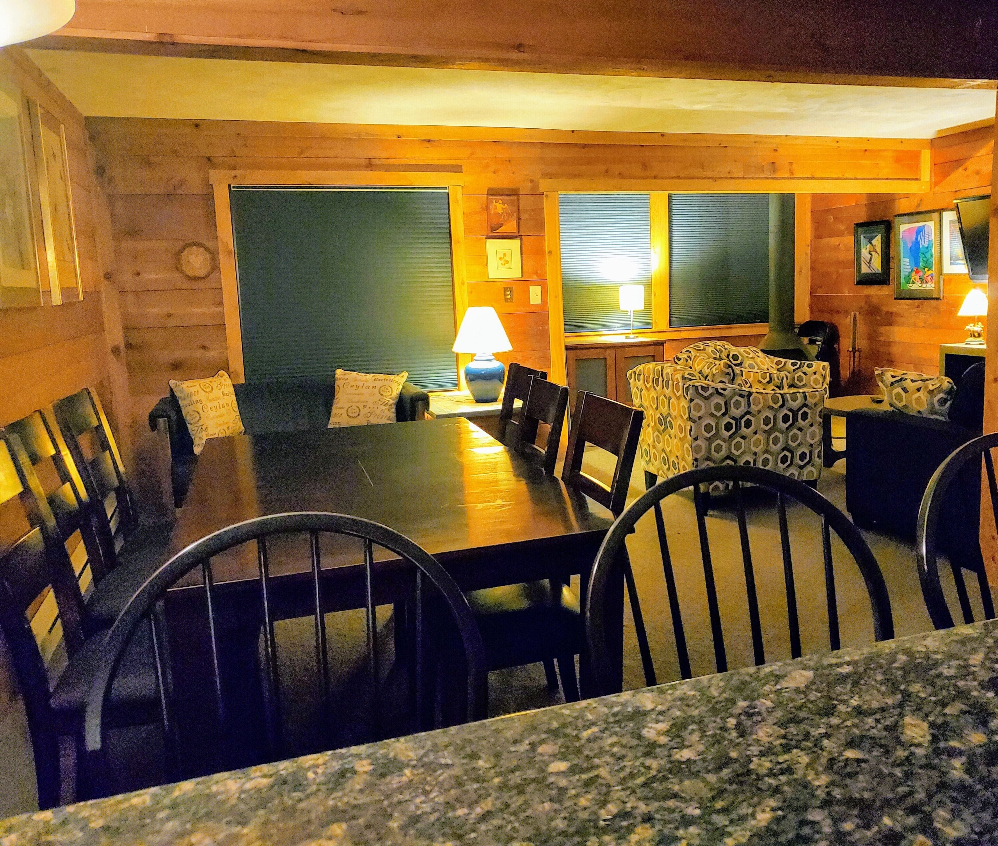 A cozy dining area with a wooden table and chairs, a patterned ottoman, and a lamp on a side table.