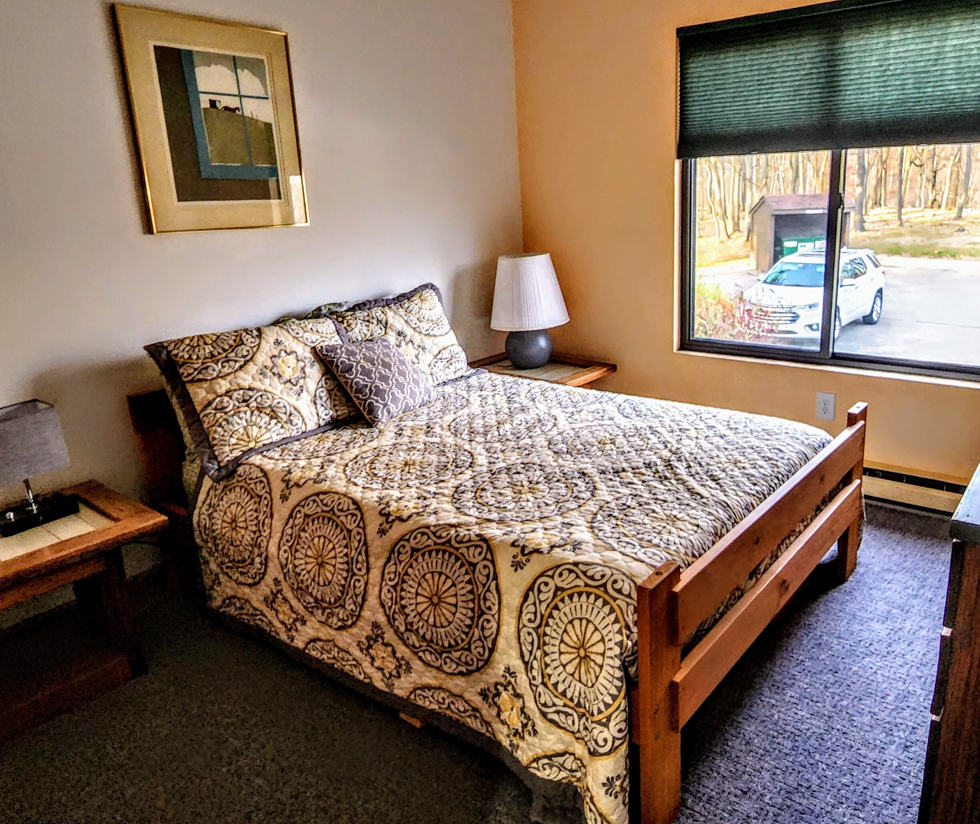 A bedroom with a patterned bedspread, a bedside lamp, a framed picture on the wall, and a window with a view of a parked car.