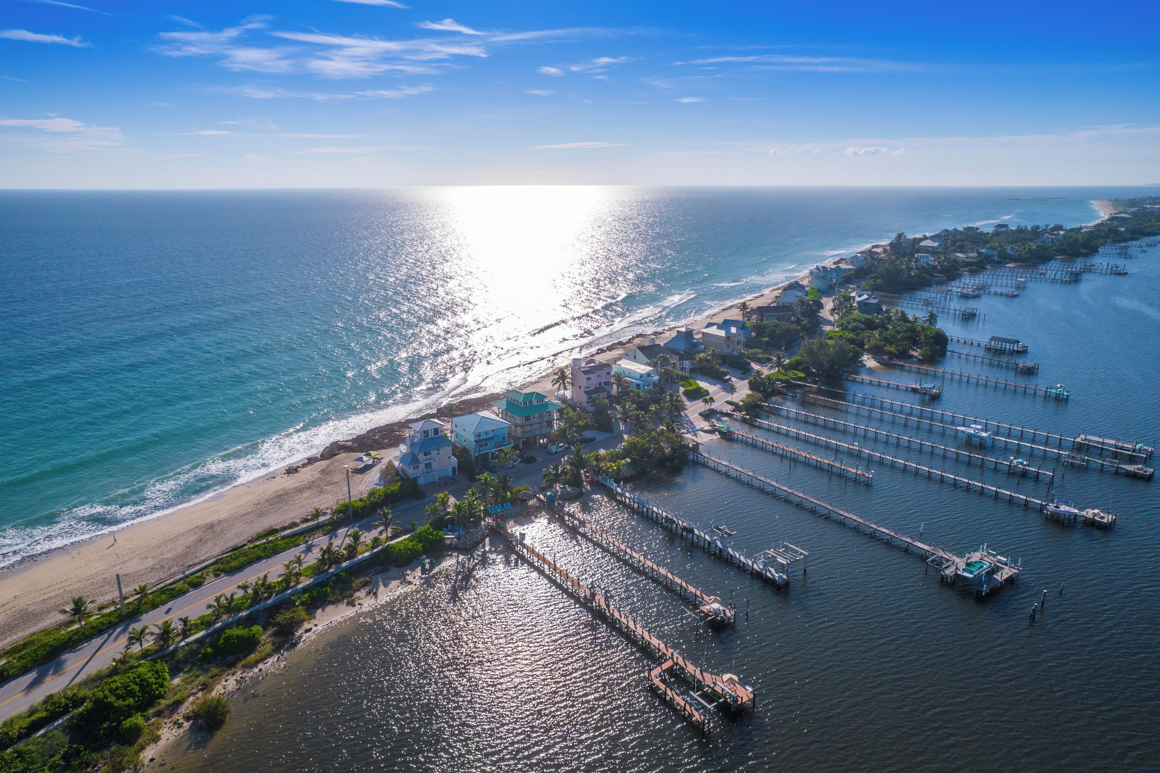 An aerial view of a coastal area with a beach, a marina with boats, and buildings with a clear view of the ocean.