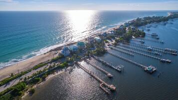 An aerial view of a coastal area with a beach, a marina with boats, and buildings with a clear view of the ocean.