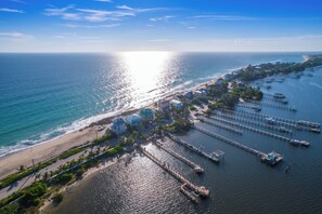 An aerial view of a coastal area with a beach, a marina with boats, and buildings with a clear view of the ocean.