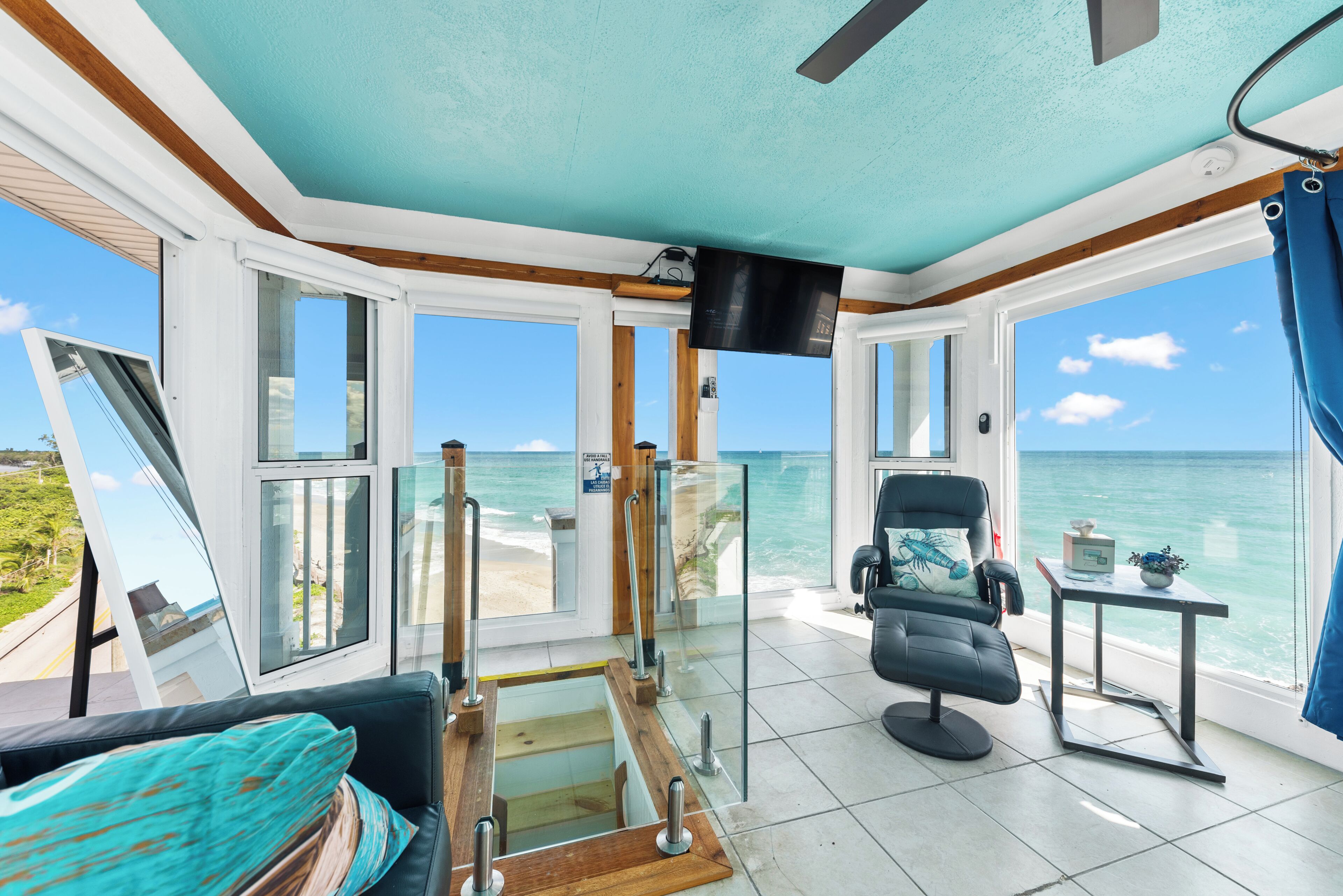 A room with a view of the ocean, a black chair, a small table with a plant, and a television mounted on the wall.