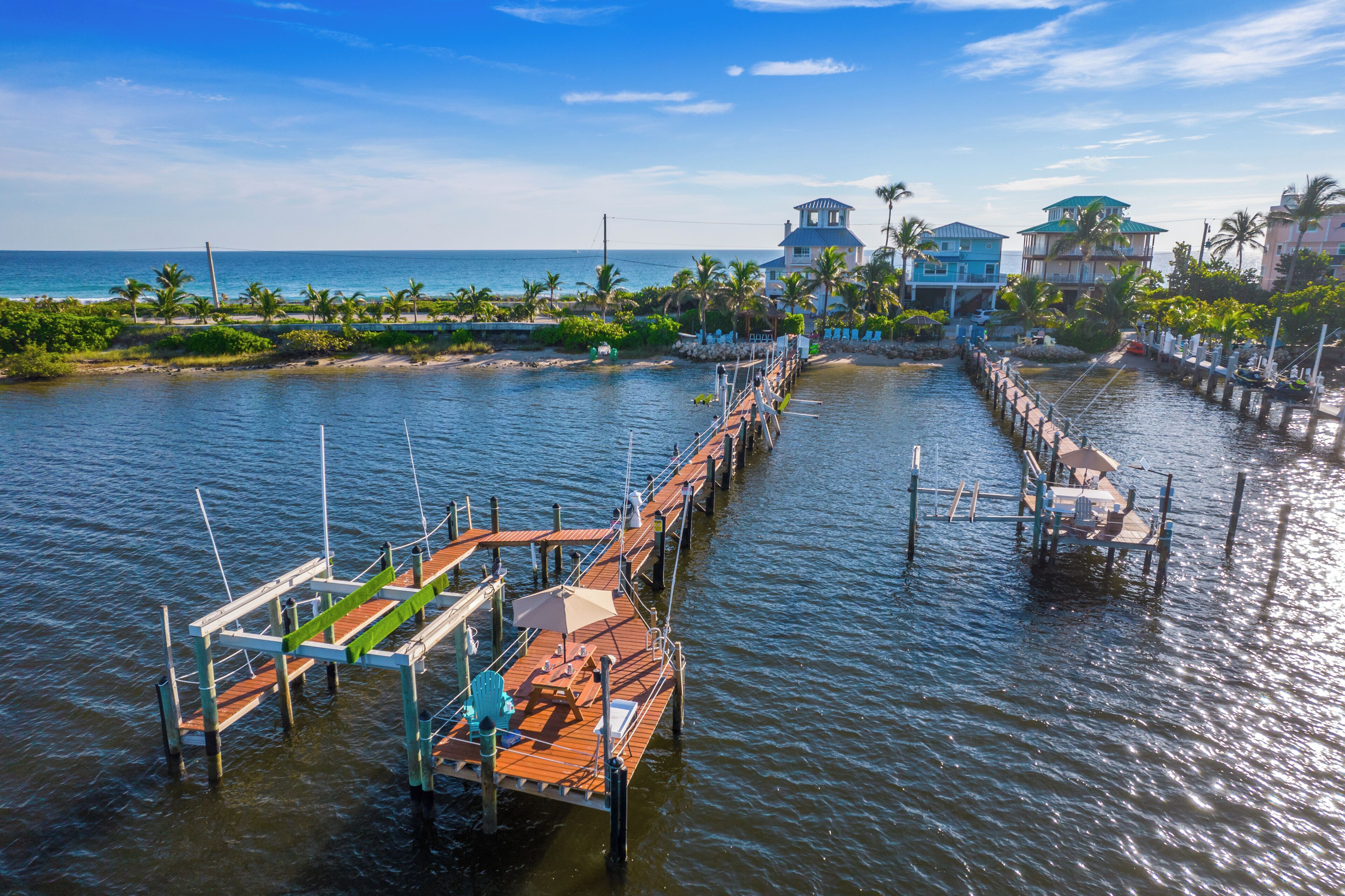 A pier extending into calm waters with a wooden deck and seating area, surrounded by tropical vegetation and buildings with blue roofs.
