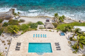 Luchtbeeld van een resort met zwembad, ligbedden, een jacuzzi en een strandgedeelte met palmbomen.