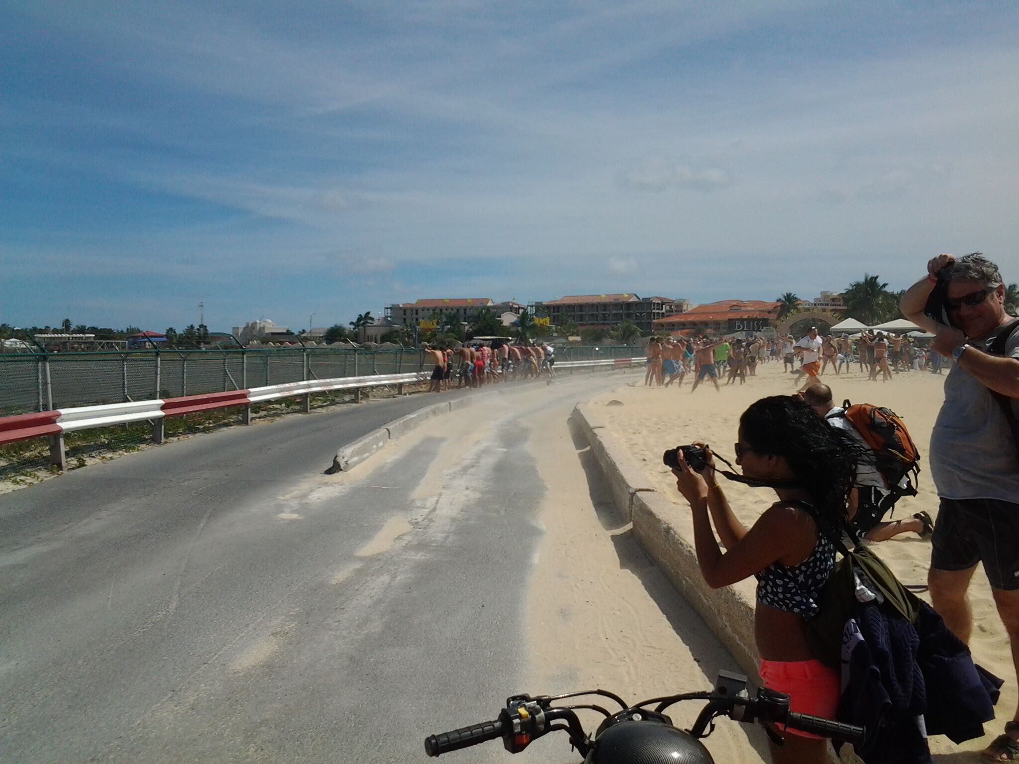 A beach scene with a road curving around it, a fence, and buildings in the background.