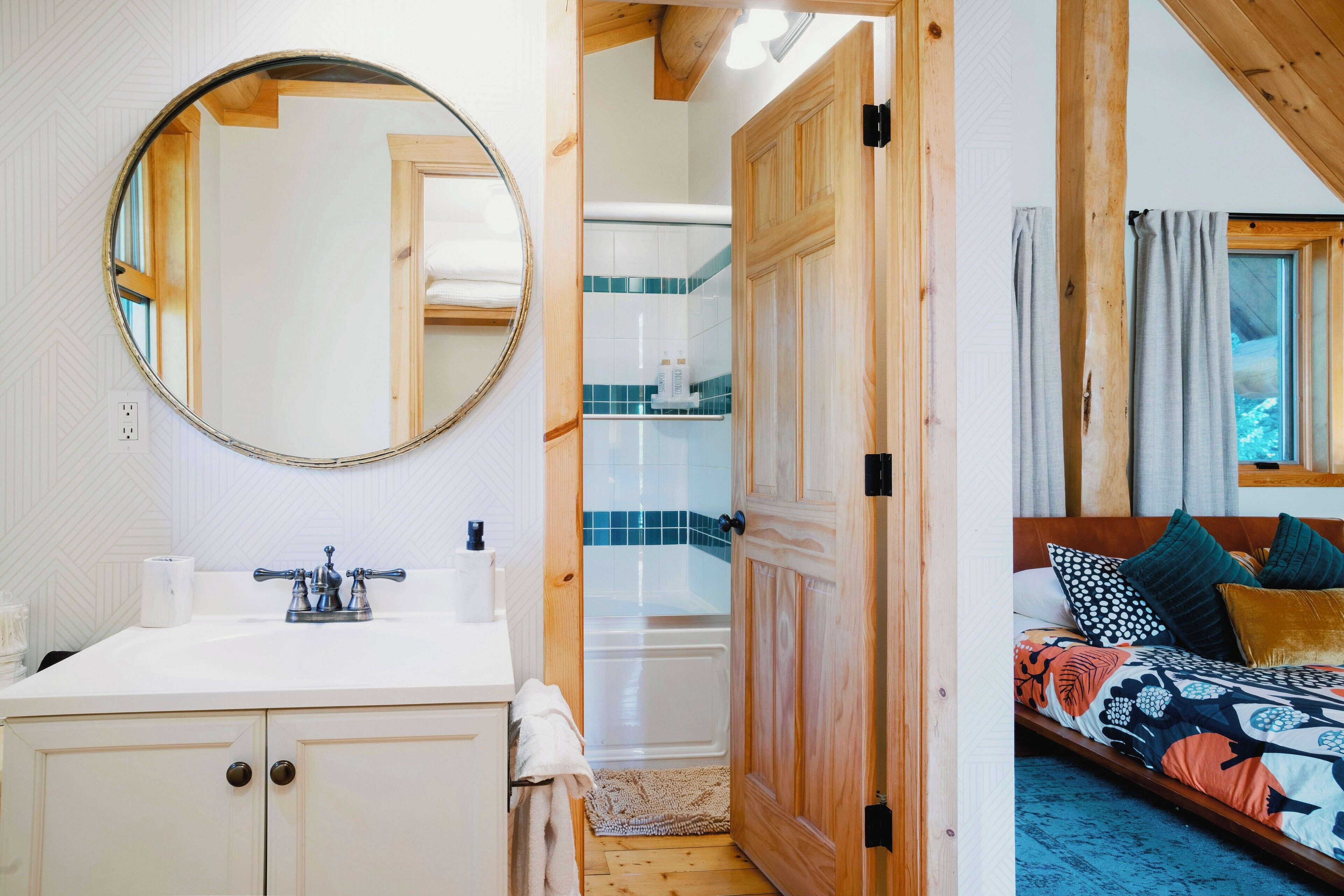 Ein Badezimmer mit weißem Waschbecken, einem Spiegel und einer Holztür, die zu einem Schlafzimmer mit Bett und Fenster führt.