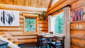 A log cabin interior with a dining area, wooden walls, a window with curtains, and a painting on the wall.
