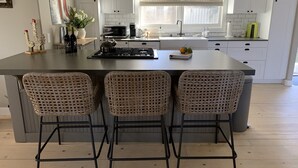 Una cocina moderna con una isla central, tres taburetes de mimbre, armarios blancos y un amplio ventanal que proporciona luz natural.