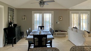 Un comedor con una mesa de madera, sillas y un ventilador de techo. Se observa un área de cocina con un mueble oscuro y un espejo. En el fondo, se ve un sofá blanco.