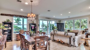 A spacious living area with a large dining table, wooden chairs, and a chandelier. There are bookshelves, a sofa, and a coffee table. - ☀︎🏝⛱︎ Beachfront Elegance⛱︎🏝☀︎  
[2026 Spring + Summer Weeks Available Now!] (North Captiva)