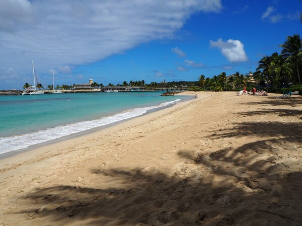 A sandy beach with clear turquoise water, a few boats, and a distant shoreline with buildings and palm trees.