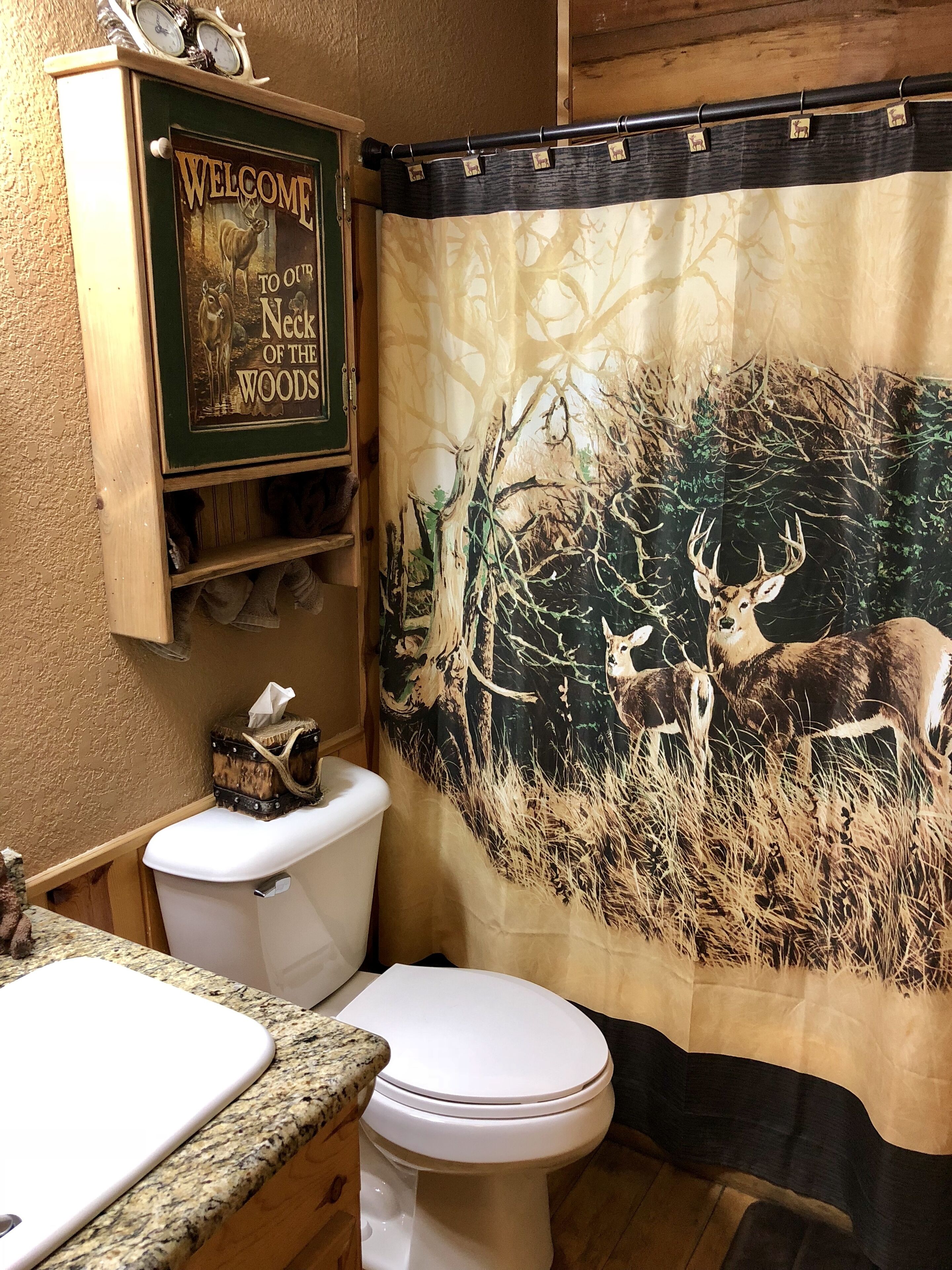 Un baño con cortina de ducha temática de ciervo, un inodoro, un lavabo con encimera de granito y un armario de madera con un letrero que dice 'Welcome to our Neck of the Woods'.