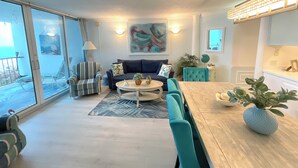A spacious living room with a large window, a dining area with a wooden table, and a kitchen area with a hanging light fixture.