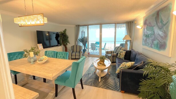 A modern living room with a dining area, a large TV, and a balcony with a view of the sea.
