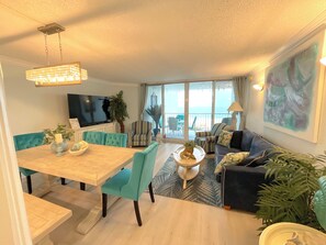 Ein modernes Wohnzimmer mit Essbereich, einem großen Fernseher und einem Balkon mit Meerblick.