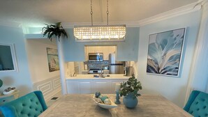 A dining area with a wooden table, teal upholstered chairs, a chandelier, and a kitchen in the background.