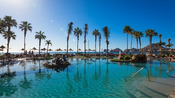 Ein klares Schwimmbecken mit Liegestühlen und Sonnenschirmen, umgeben von Palmen und Blick auf den Ozean.