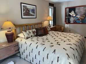 A bedroom with a wooden headboard, a bed with a patterned duvet, a nightstand with a lamp, and a framed picture on the wall.