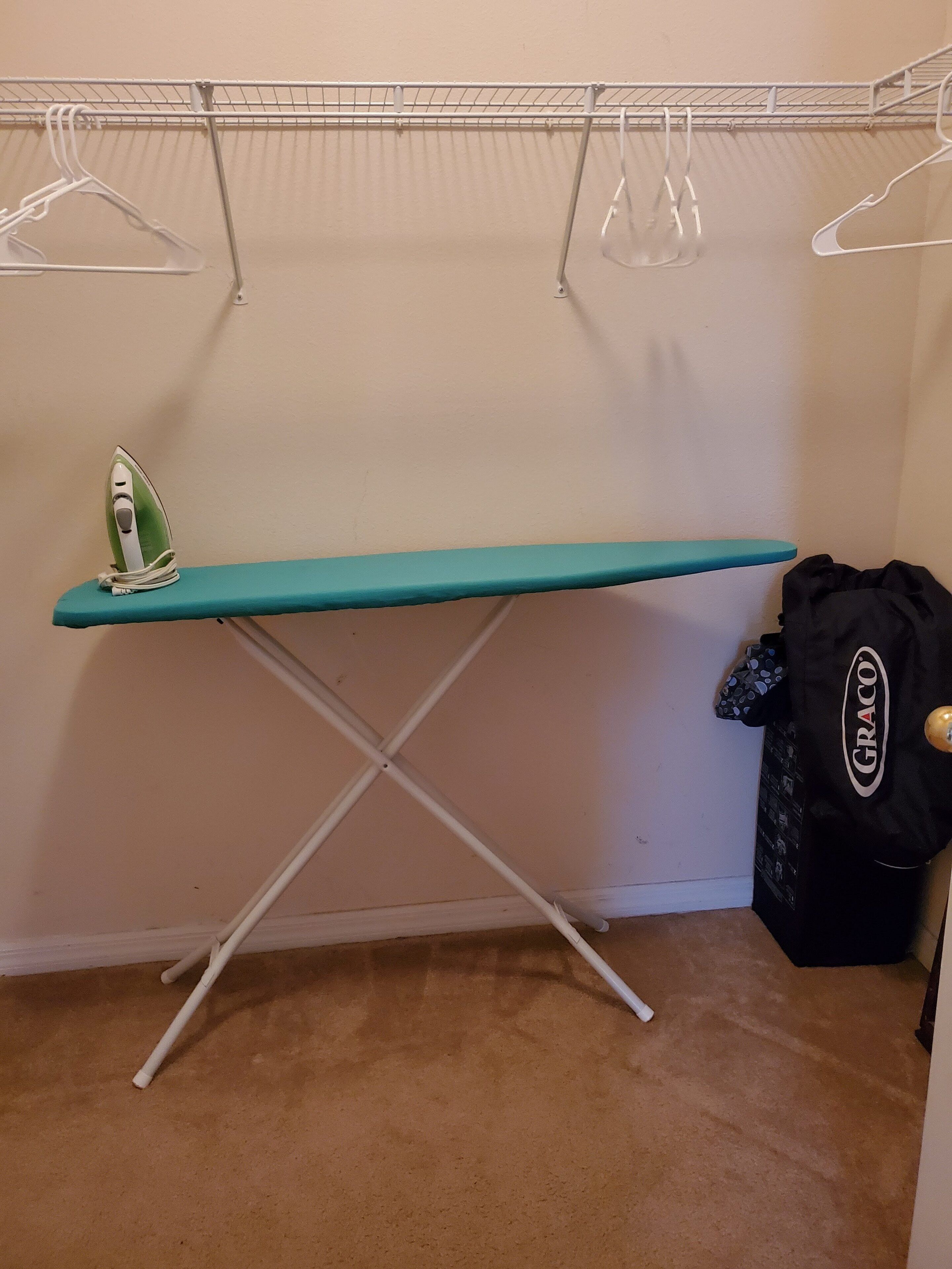 Una habitación con una tabla de planchar color turquesa, una bolsa Graco y otra tabla de planchar blanca en la esquina.