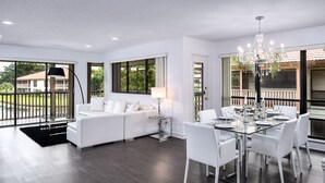 A spacious living room with a white sofa, a dining table set for six, and large windows offering a view of the outdoors.
