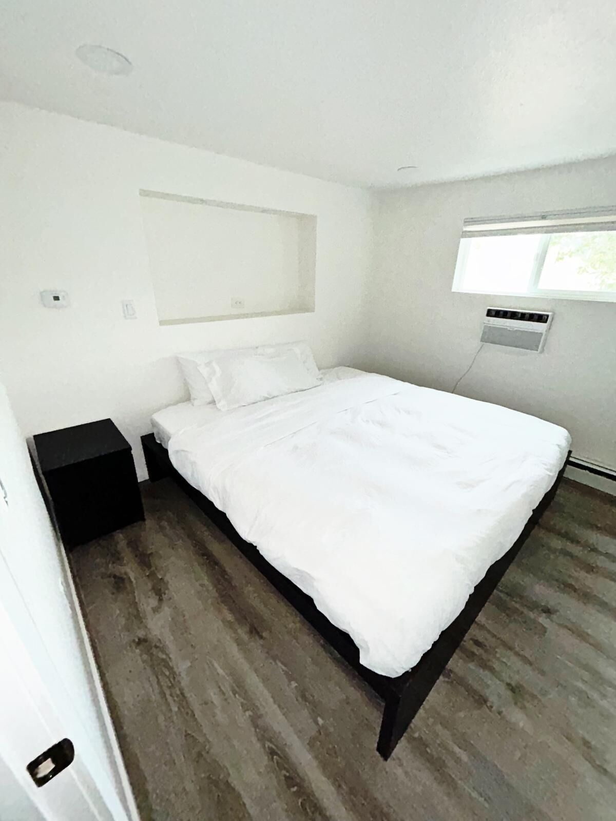 Une chambre à coucher comprenant un lit, une table de chevet, une fenêtre et un climatiseur fixé au mur.