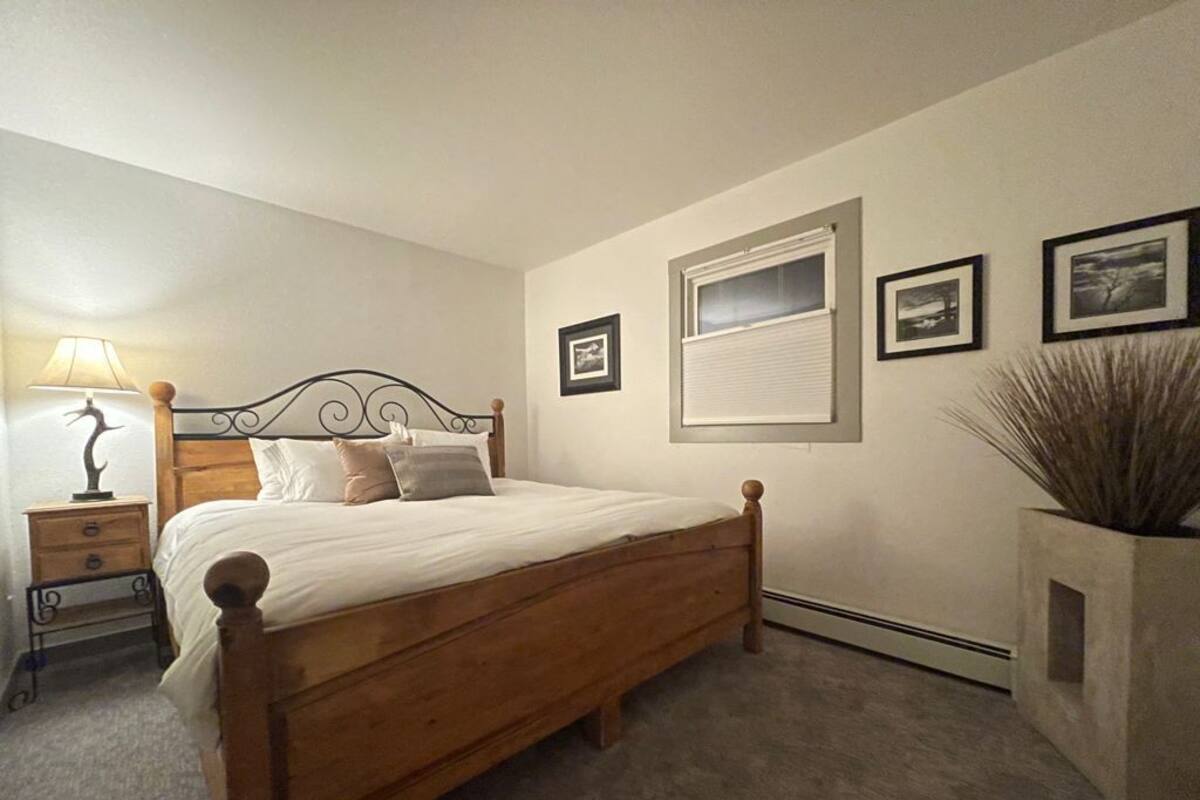 Un dormitorio con una cama de madera, una mesita de noche con lámpara, una ventana con persianas e imágenes enmarcadas en la pared.