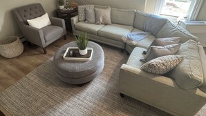 A living room with a grey armchair, a beige sofa, and a round ottoman with a plant. There is a small table with a lamp and a potted plant.