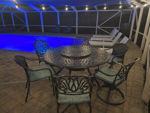 An outdoor patio with a round metal table and chairs, a pool, and a covered seating area.