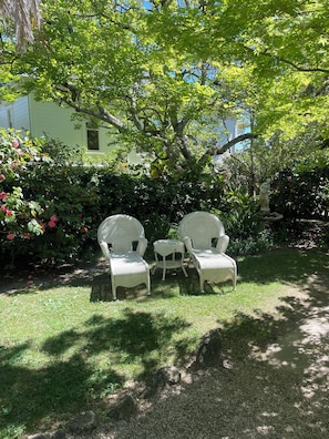 Two wicker chairs and a small table in a garden with a house in the background.