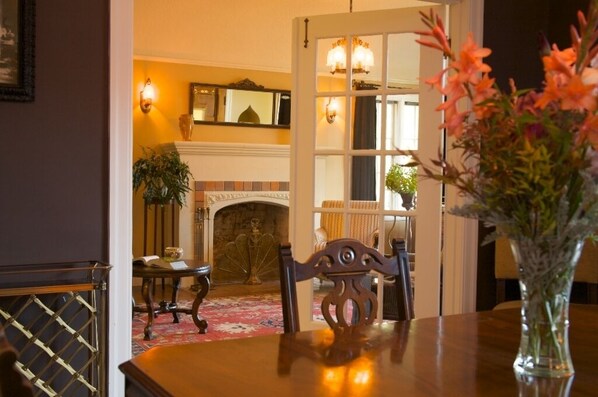 A dining area with a wooden table, chairs, and a vase of flowers. There is a fireplace in the background with a mantelpiece and a mirror above it.