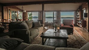 A cozy living room with a large window overlooking a lake and hills. There is a sofa, a chair, and a coffee table. - Lakeside Lodge - 15 ft From Lake, Dock, Kayaks/Canoes/SUPs, Pet Friendly! (Rangeley)