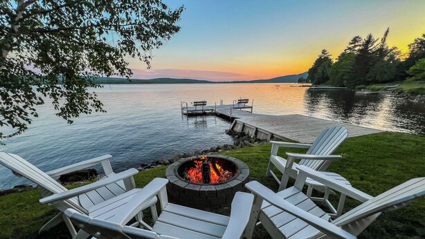 A lakeside deck with Adirondack chairs, a fire pit, and a dock extending into the water. - Lakeside Lodge - 15 ft From Lake, Dock, Kayaks/Canoes/SUPs, Pet Friendly! (Rangeley)
