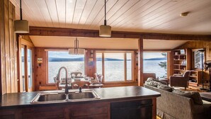 A cabin with a kitchen featuring a large island with a sink, a dining area with a table set for four, and a seating area with a sofa and armchairs. Large windows provide views of a lake and mountains. - Lakeside Lodge - 15 ft From Lake, Dock, Kayaks/Canoes/SUPs, Pet Friendly! (Rangeley)