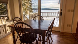 A dining area with a wooden table and chairs, a view of a lake and mountains through a glass door, and a wooden deck outside. - Lakeside Lodge - 15 ft From Lake, Dock, Kayaks/Canoes/SUPs, Pet Friendly! (Rangeley)