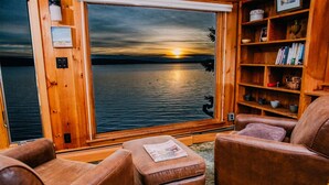 A cozy cabin interior with a large window overlooking a sunset over a body of water, leather furniture, and a wooden bookshelf. - Lakeside Lodge - 15 ft From Lake, Dock, Kayaks/Canoes/SUPs, Pet Friendly! (Rangeley)