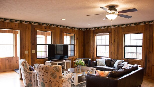 Un salon avec des murs en bois, un ventilateur de plafond, un téléviseur à écran plat, un canapé, deux fauteuils à motifs et une table basse sur laquelle est posé un vase.