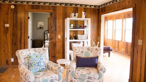 Une pièce chaleureuse aux murs en bois, avec deux fauteuils à motifs, une étagère blanche et une vue sur l’extérieur par une porte ouverte.