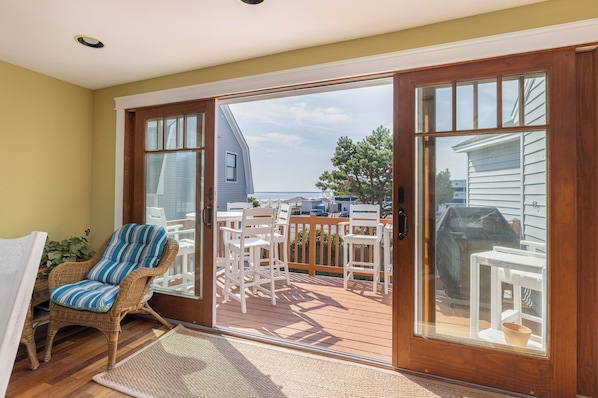 A covered deck with white chairs and a wicker chair, a wooden door, and a view of the sea and buildings.