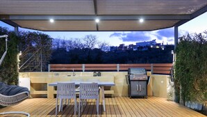 Eine Außenpatio mit Holzdeck, einem Tisch mit Stühlen, einem Grill und einem überdachten Bereich mit Beleuchtung.
